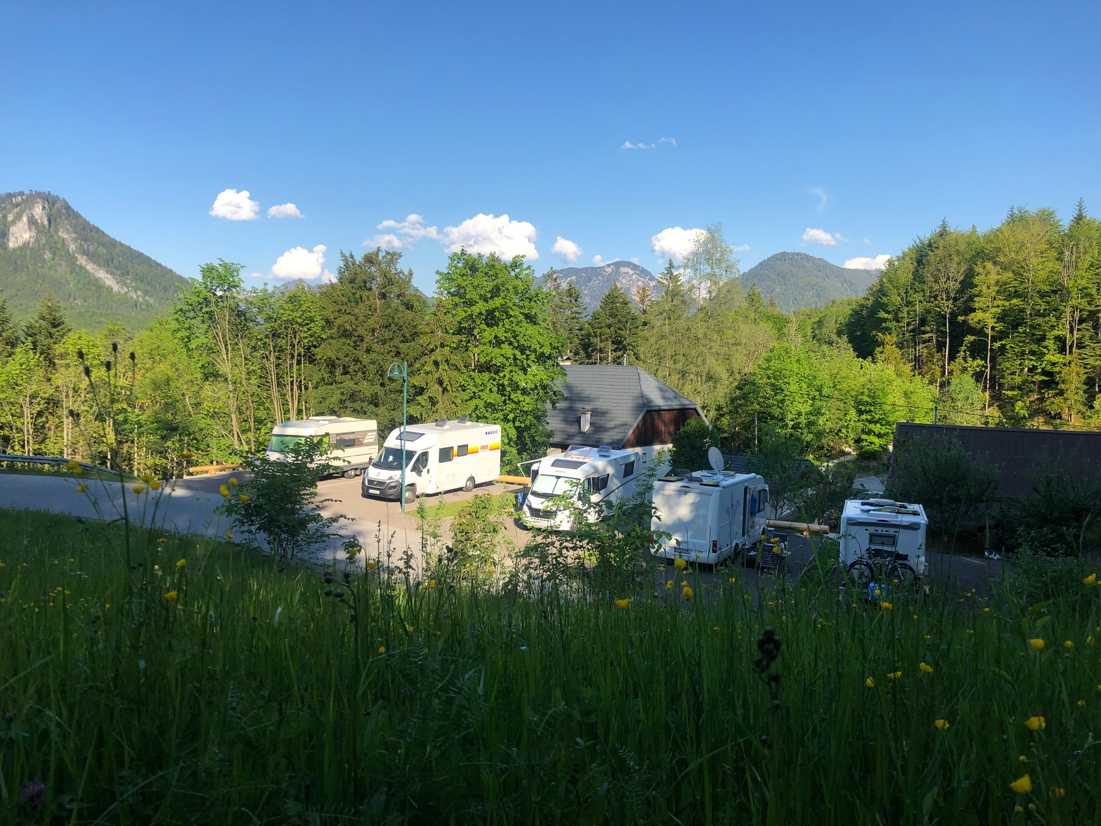 Mehrere Wohnmobile parken auf einem Stellplatz inmitten einer grünen Berglandschaft unter blauem Himmel.