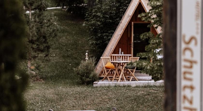 Gemütliche A-Rahmen-Holzhütte mit Sitzgruppe auf einer Wiese, umgeben von Bäumen und Sträuchern.