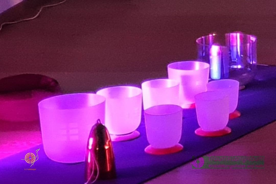 Eine Reihe mattierter Kristall-Klangschalen, die in violettem Licht auf einer dunklen Matte leuchten.