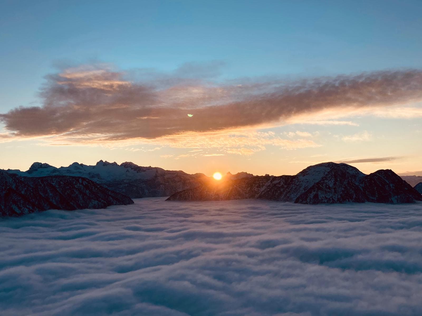 Die Sonne sinkt hinter schneebedeckte Berge über einem weiten, wellenförmigen Meer aus weißen Wolken.