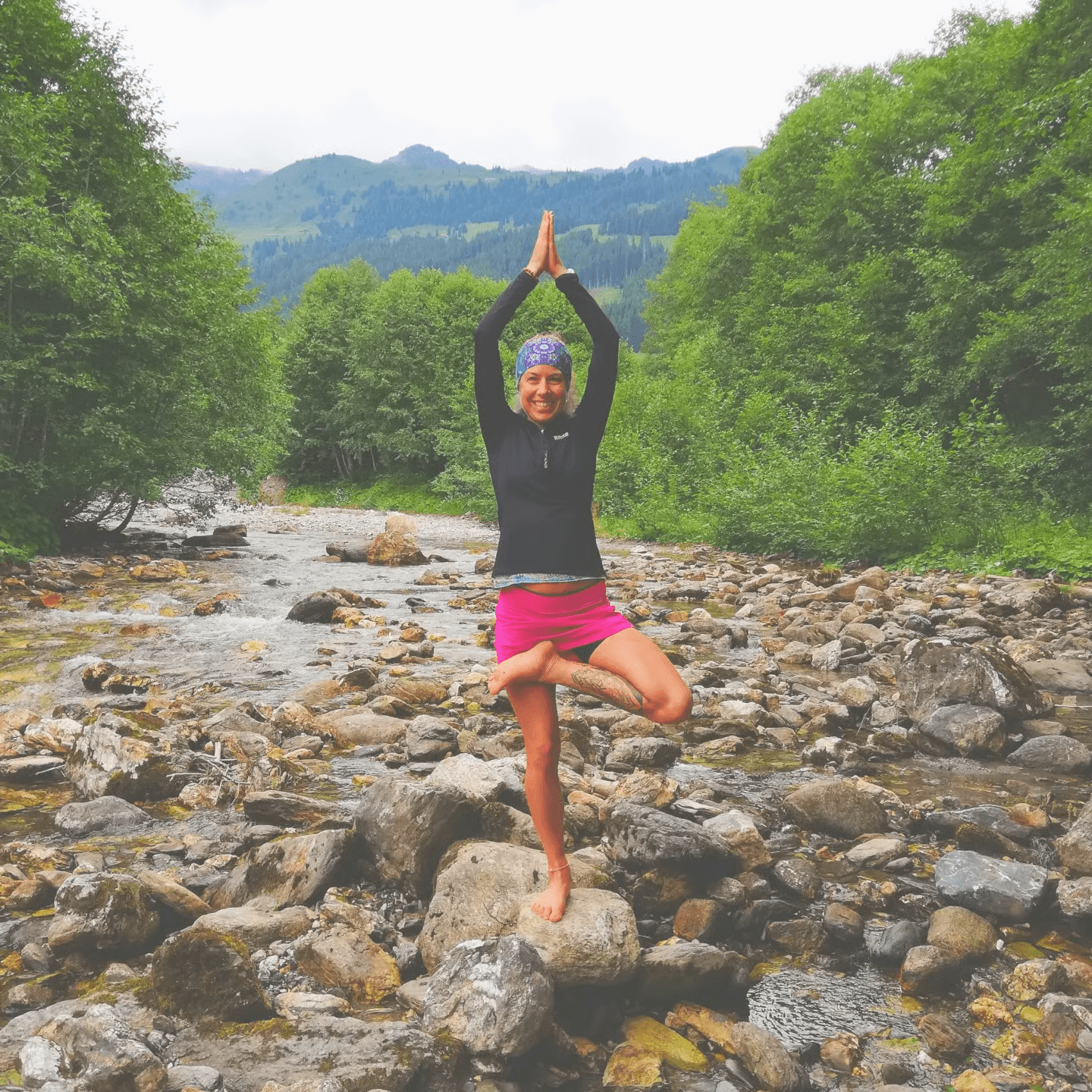Eine Frau praktiziert Yoga auf einem Felsen in einem Gebirgsbach vor bewaldeten Bergen.