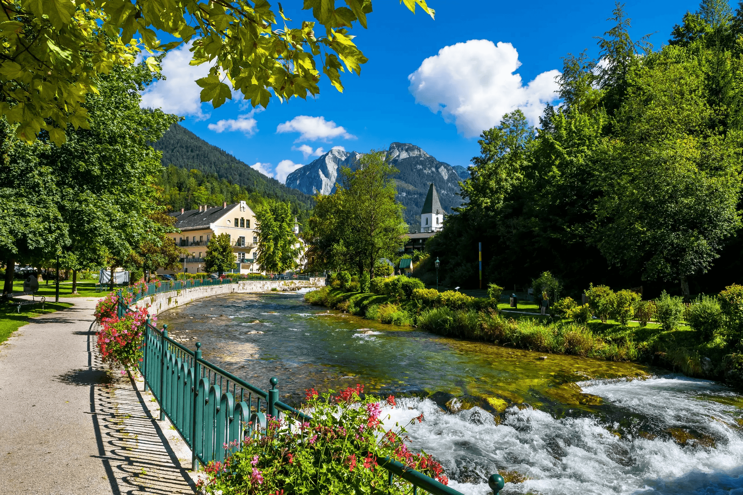 Malerischer Flusslauf in Bad Ischl mit blumengeschmücktem Uferweg vor einer beeindruckenden alpinen Bergkulisse.